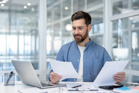 Business man with a beard smiling while reviewing financial documents, managing paperwork, and using a laptop at his desk in a modern corporate office, symbolizing planning and success