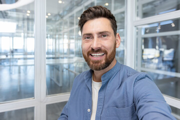 Happy bearded man smiling during a video call or digital conference, capturing a selfie portrait in a modern glass office while communicating effectively