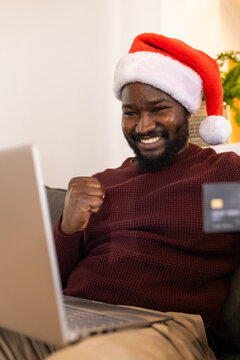 African American man sitting at home on sofa wearing Santa hat holding credit card, using laptop