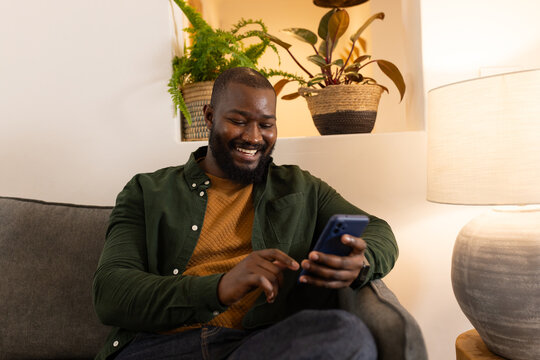 African American man sitting on gray sofa in living room corner using smartphone with woven baskets