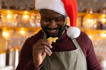 African American man holding holiday-shaped cookie near mouth with olive apron, Santa hat in cafe