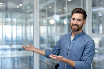 Smiling bearded man gesturing with open hands, welcoming to a sleek, modern office interior with glass partitions, representing hospitality and business invitation