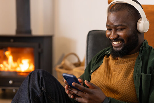African American man sitting at home by wood stove with headphones, holding smartphone, copy space