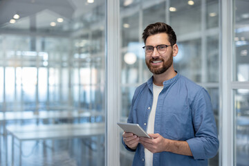 Young businessman wearing glasses and a blue shirt holding a digital tablet and smiling at the...