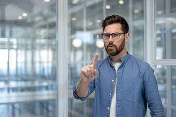 Serious male businessman wearing glasses and a blue shirt, standing in a modern office and intently pointing a finger forward, interacting with an invisible augmented reality interface