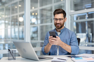 Obraz premium Smiling businessman wearing glasses and a beard sitting at a desk, looking cheerful while messaging on a smartphone, with a laptop and documents on the contemporary office table