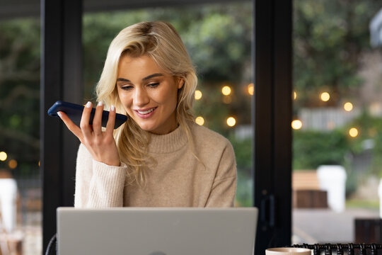 Female speaking into smartphone and viewing laptop at cafe wooden table with cup and string lights