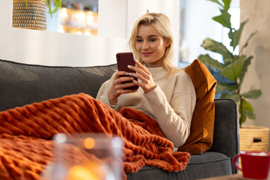 Adult woman sitting on gray sofa at home holding smartphone with orange blanket and candlelight