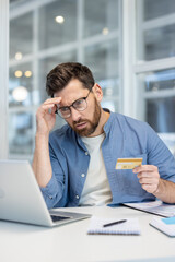 Man finding it difficult to manage debt while holding a credit card and using a laptop for online banking, feeling overwhelmed by financial problems and budget constraints