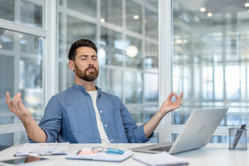 Fototapeta premium Young businessman meditating at his office desk in a mudra pose, eyes closed, finding calm and work-life balance while managing stress in a modern workplace setting