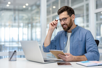 Smiling man in smart-casual attire holding glasses and a notebook while working on a laptop in a...