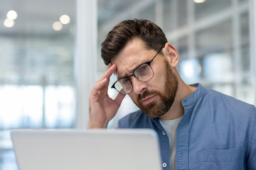 Young bearded man with glasses at a laptop, rubbing his temples and wincing from a headache while...