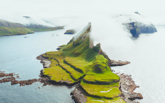 Aerial view of the dramatic, craggy island of Drangarnir, swathed in ethereal mists and verdant green slopes meeting the steely blue sea, Drangarnir, Faroe Islands.