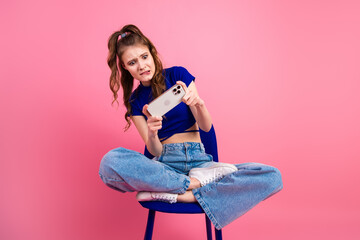 Young woman with brunette hair holding smartphone, wearing blue top and jeans, sitting casually on...