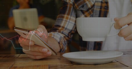 Holding smartphone and ceramic coffee cup, man sitting at coffee shop table, with saucer and laptop