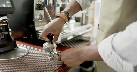 Barista wearing taupe apron tamping grounds into portafilter at coffee bar with grinder, steam wand