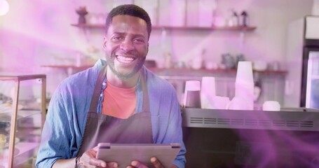 Smiling male barista wearing red shirt and dark apron, holding tablet at cafe, with coffee machine