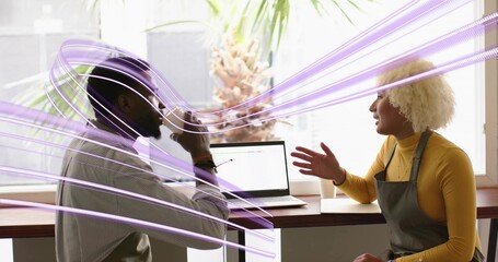 Sitting barista wearing gray apron and customer chatting at coffee shop counter, with laptop, mugs