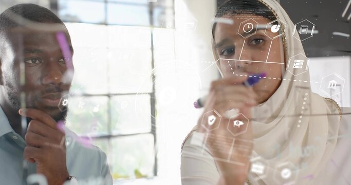 Collaborating man in shirt and woman in hijab planning on glass board at office, digital icons