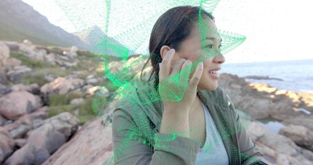 Adjusting earbud Asian woman on rocky outcrop by ocean, with green AR mesh overlay, copy space