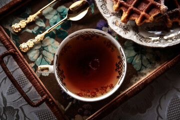 Elegant cup of tea on a floral tray with golden spoons and waffles on a vintage plate, creating a cozy and classic breakfast atmosphere.