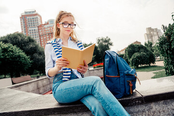 Beautiful woman reading a book outdoors on a sunny urban day, sitting stylishly near her blue...