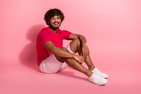 Smiling young man in stylish casual attire seated against a vibrant pink background, expressing leisure and positivity