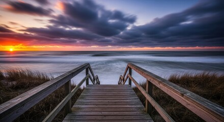 Obraz premium Wooden boardwalk leading down to the ocean at sunset with dramatic clouds and waves
