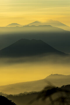 View of majestic mountain peaks pierce the hazy sunrise, casting long shadows across the undulating landscape, creating a serene atmosphere, Bromo, East Java, Indonesia.