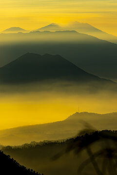 View of layered, misty mountains rising into a golden sky, with soft light creating depth and a sense of serene grandeur, Bromo, East Java, Indonesia.