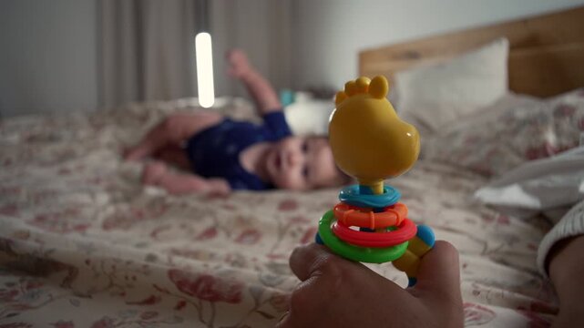 Hand spinning colorful baby toy while infant lies on bed watching, playful interaction stimulating sensory development, curiosity, and focus during early learning at home
