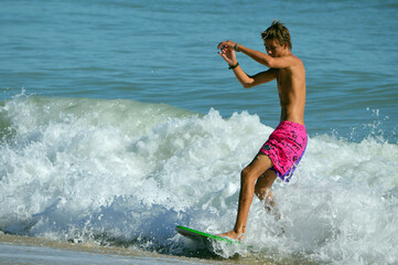 Fototapeta premium A teenage skimboarder glides across the surf, arms raised and leaning back as waves crash all around him on a sunny Florida beach.