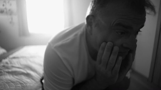 Man sitting on bed covering face with both hands, expressing emotional pain, exhaustion, and despair during quiet moment of sadness and mental struggle in black and white