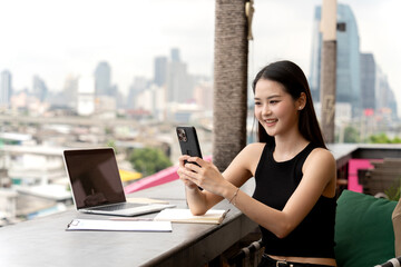 Asian adult woman uses smartphone rooftop cafe city view digital nomad online connection happy remote worker modern lifestyle global communication laptop use.