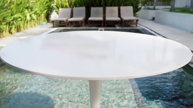Clean white table in focus with a blurred background of pool and tropical greenery. Minimal composition with slow motion camera movement. Ideal for mockups and lifestyle use.