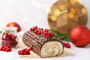 Delicious Yule log (traditional Christmas cake) with redcurrant and decor on white table against blurred lights, closeup. Bokeh effect