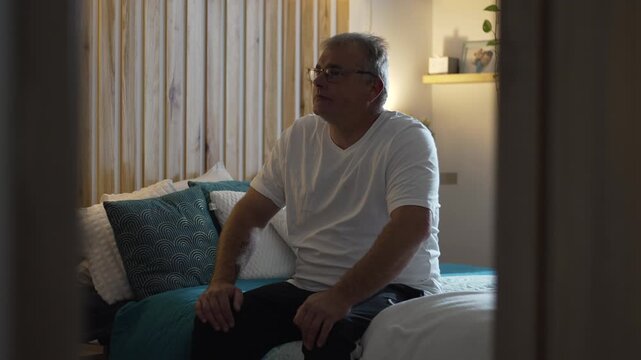 Middle aged man sitting still on bed in quiet room appearing lost and purposeless struggling with emptiness and lack of direction in a moment of silent existential fatigue
