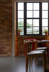 A cozy indoor space with wooden chairs, a brick wall, potted plants on a windowsill, and a large window letting in natural light.
