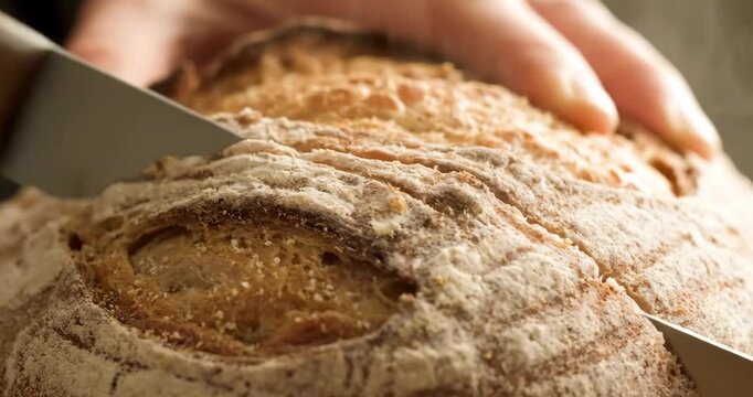 4K macro image. A serrated knife gently slices through a crusty, golden-brown loaf of bread. The texture of the bread as the crust breaks and reveals the soft, steamy interior.