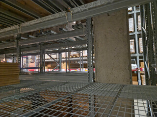 Interior low-angle view of a large, multi-level warehouse storage system, featuring steel pallet racking, a concrete support column, and a metal grid floor on the upper level.