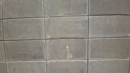Close-up, and slightly angled view of a simple, unpainted grey concrete block wall or textured pavement, displaying a pattern of rectangular blocks separated by thin grout lines.
