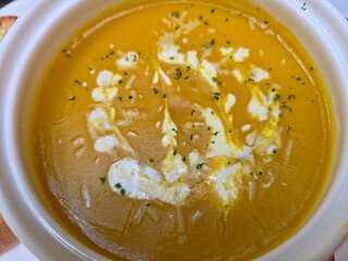 A top-down close-up of a bowl of creamy, rich orange pumpkin soup garnished with a drizzle of white cream or yogurt and a sprinkle of fresh herbs like parsley.