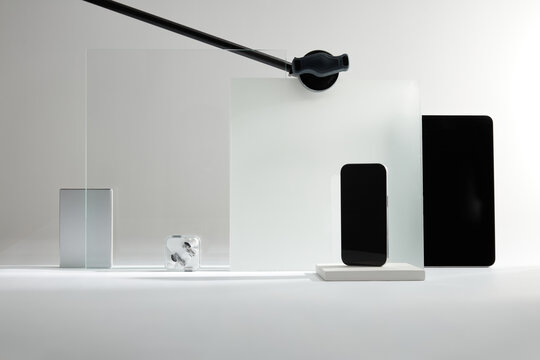 Mockup of Modern Consumer Electronics: Smartphone, Tablet, and Earbuds Displayed with Translucent Panels in a Minimalist Studio Setting.