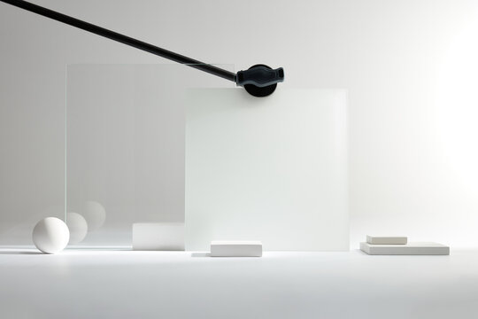 Abstract geometric composition featuring a white sphere, rectangular blocks, and translucent glass panels held by a black mechanical arm in a bright, minimalist studio environment.