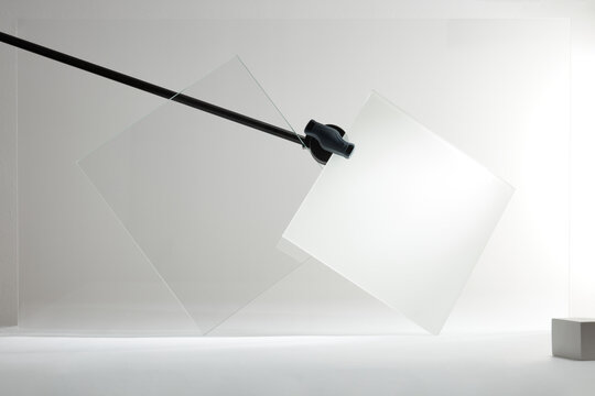 Abstract display of two glass panels (clear and frosted) held by a black mounting rod, showcased in a minimalist, high-key studio environment emphasizing geometric forms and material technology.