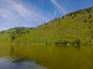 Scenic view of lush green hillside reflecting in calm river under clear blue sky