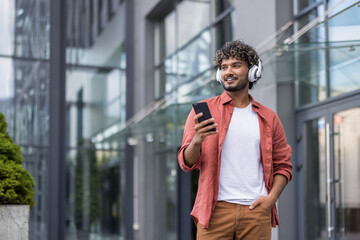 Photo of a young smiling Indian man wearing headphones walking around the city, listening to music and using his phone