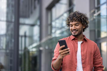 Close-up photo of a young Indian man standing on the street with a smile and using a mobile phone