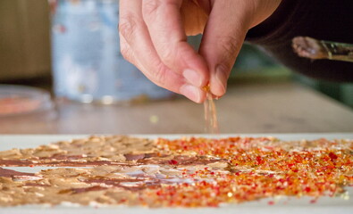 A hand sprinkles fine red glass crumbs or sand onto a colorful glass art project