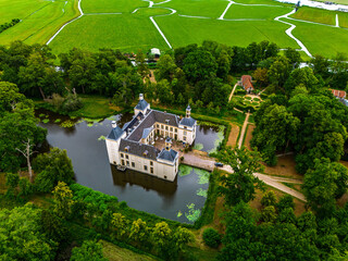 Aerial view of historic castle symmetrical twin towers and central hall framed by forest, moats, and tree lined path, with open fields and residential zone beyond under warm sunrise sky.
